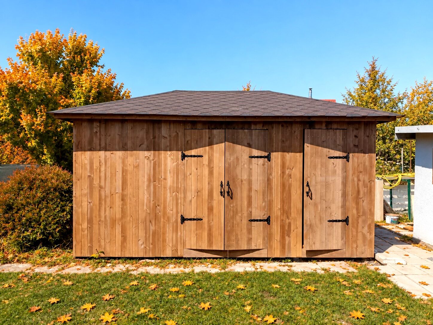 A wooden shed with double doors surrounded by autumn foliage and fallen leaves.